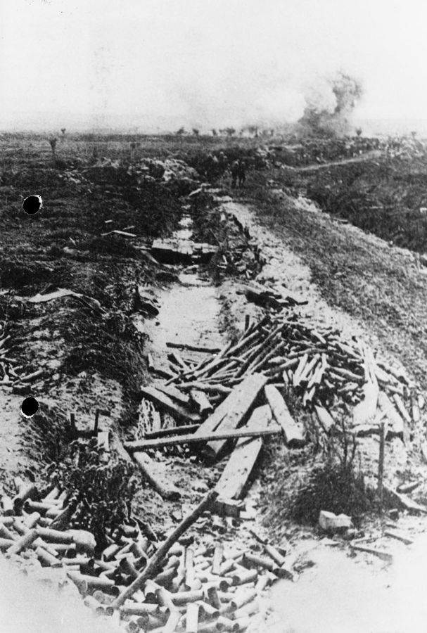 A pile of 18-pounder shell cases show that a gun crew are to the left of the photo, out of sight. An artillery shell explodes in the background. Ypres Salient.